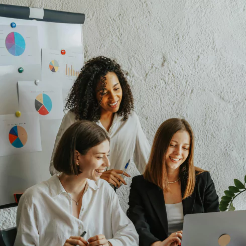 Three women in business attire collaborate at a table, focused on a laptop